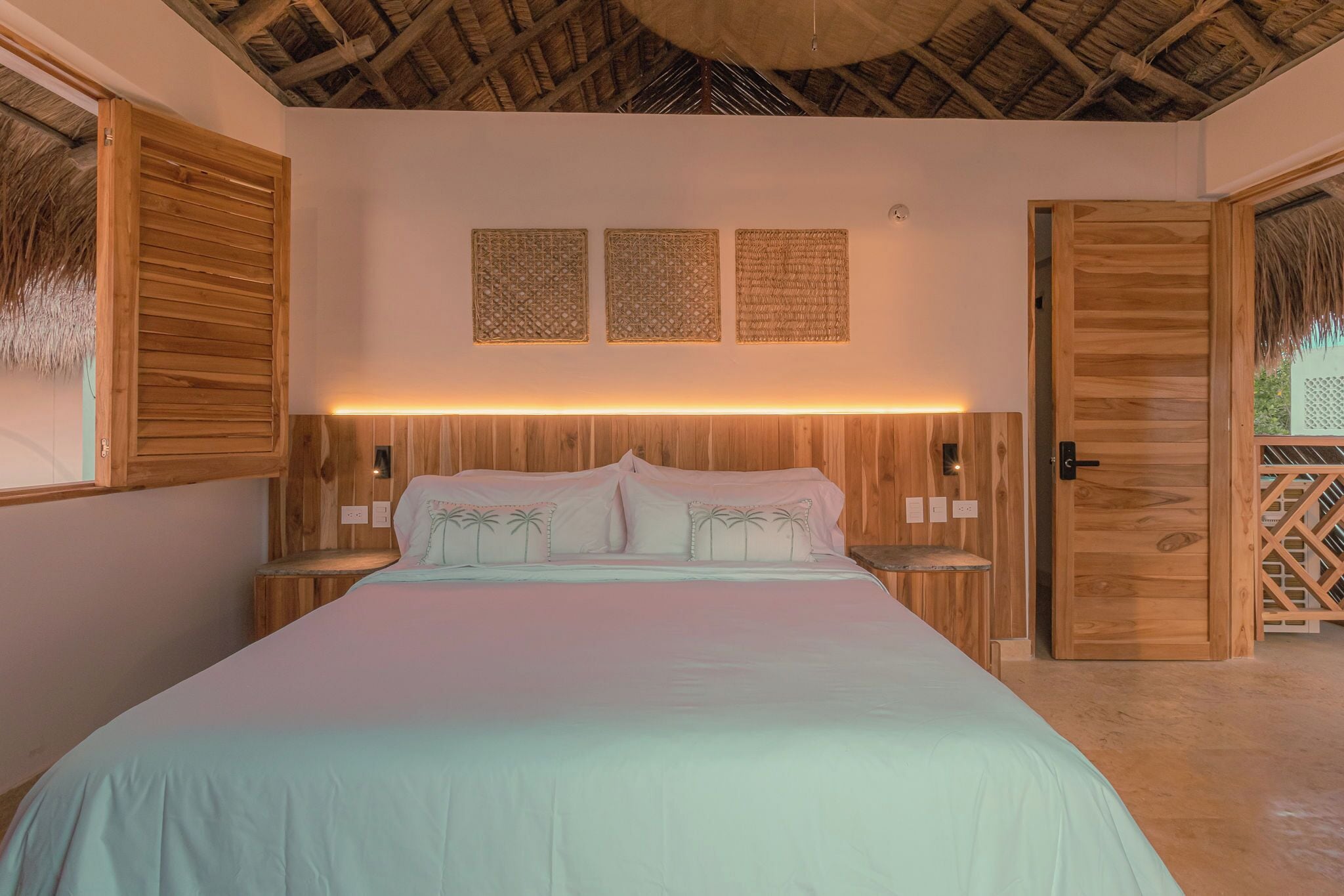 Un dormitorio con cabecera de madera, cama con ropa blanca, dos luces fijadas a la pared y una puerta con textura tejida.