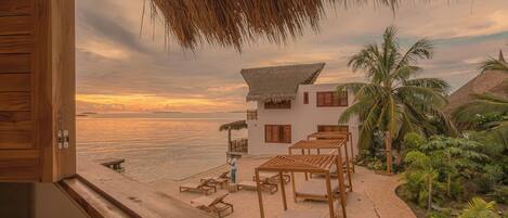 A wooden deck with lounge chairs and a thatched roof structure overlooking a beach with a white building and palm trees.