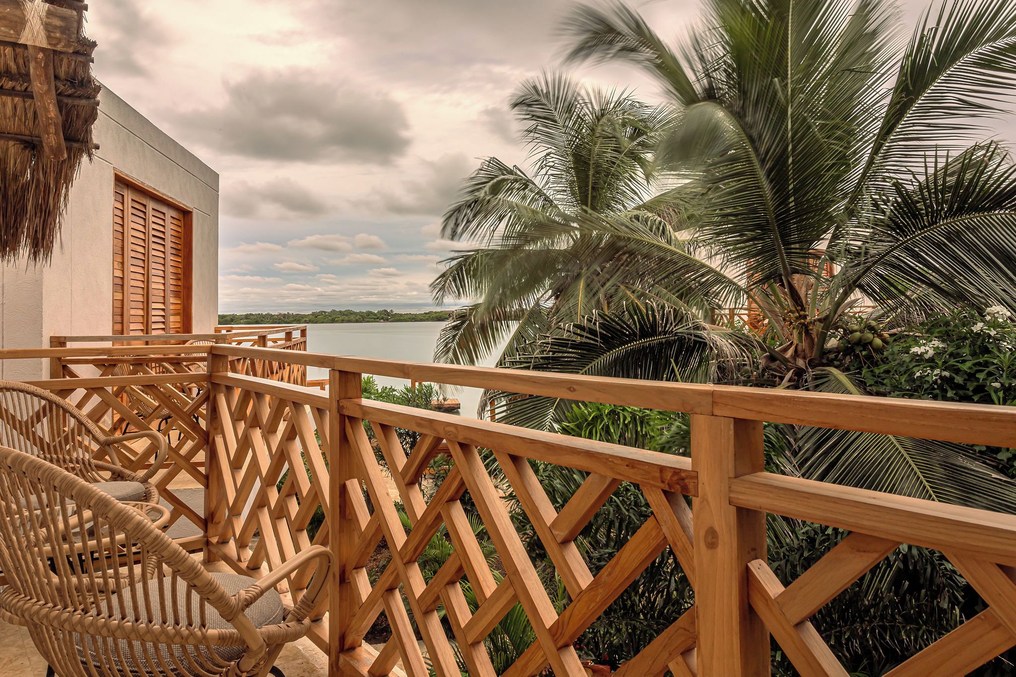 A wooden balcony with wicker chairs, a thatched roof, and a view of a lake and palm trees.