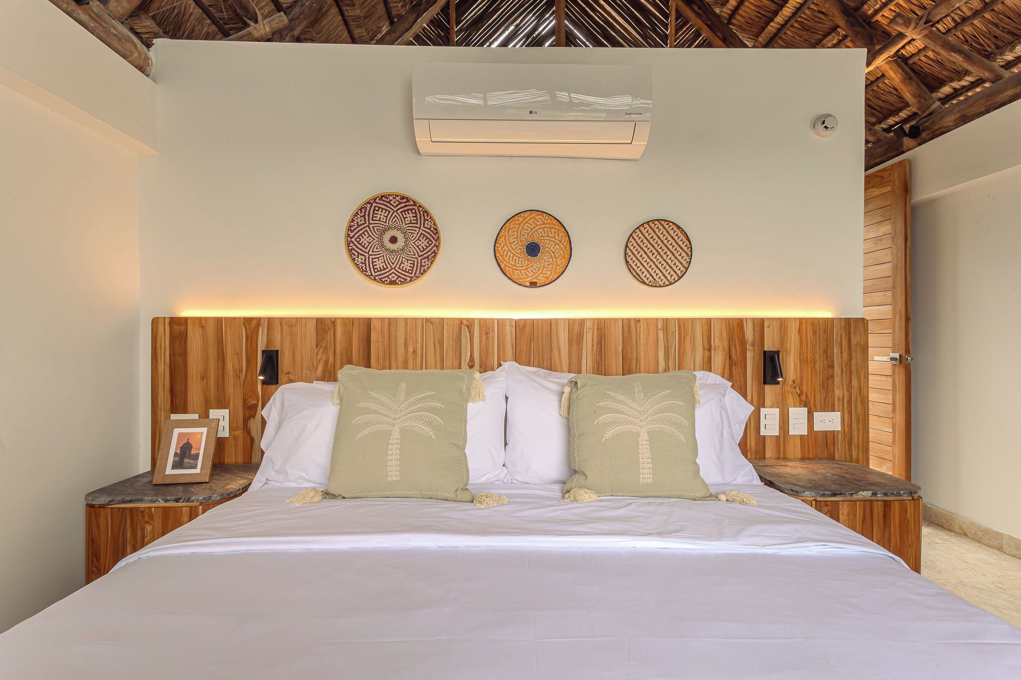 Una cama bien tendida con almohadones de hojas de palma, un cabecero de madera y un aire acondicionado de pared.