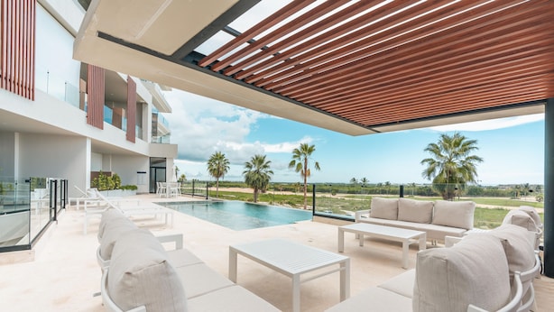 Un área lounge moderna al aire libre con piscina, mobiliario blanco y vistas a palmeras y un paisaje distante.
