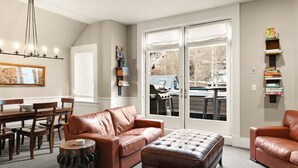 A living room with a brown leather sofa, a wooden coffee table, a dining area with a wooden table and chairs, and a large window with a view of a backyard grill and trees. - Pacific Place: Mountains On Main (Telluride)