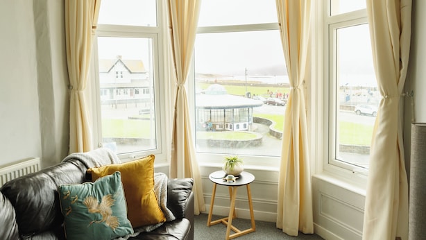 Una habitación acogedora con un sofá de cuero, una mesita redonda con una planta y ventanas grandes con cortinas.