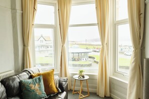 A cozy room with a leather sofa, a small round table with a plant, and large windows with curtains. - Seaside Escape: Peaceful 2BR in Picturesque Wales (Llandudno)