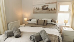 Una cama bien hecha con toallas enrolladas, un cuadro de un barco en la pared, una ventana con cortinas y una lámpara de noche.