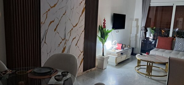 A modern living room with a marble-patterned wall, a flat-screen TV mounted on the wall, a sofa with patterned cushions, and a dining area with a table and chairs.