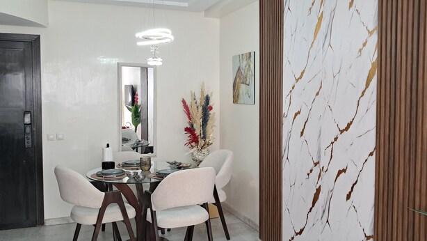Un comedor moderno con una mesa de vidrio, sillas blancas, un jarrón decorativo y una pared con estampado de mármol.
