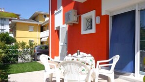 A patio with a table and chairs, a car parked nearby, and a building with a red wall and a blue door. - Apartment in Caorle Near Adriatic Beaches (Caorle)