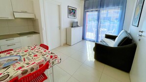 A compact living space with a kitchenette, a dining area with a table and chairs, a sofa, and a large window with curtains. - Apartment in Caorle Near Adriatic Beaches (Caorle)