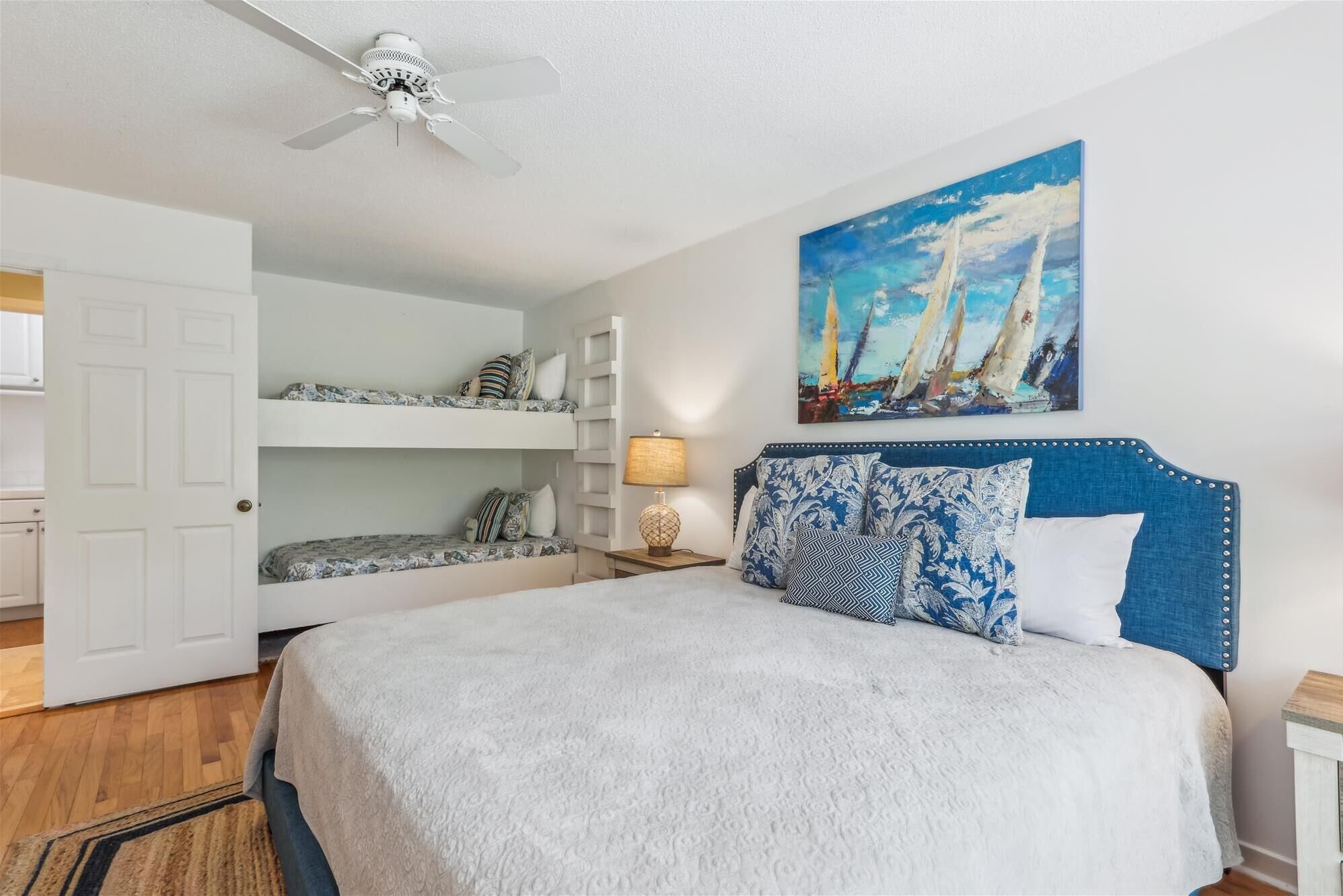 A bedroom with a bunk bed, a bed with a blue headboard, a painting of sailboats, and a ceiling fan.