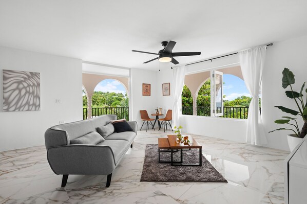 A modern living room with a grey sofa, a wooden coffee table, and a ceiling fan. There are two chairs and a small table in the background.