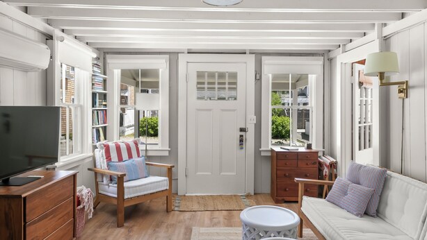 A room with a wooden floor, white walls, a white sofa, a striped chair, a TV, a lamp, and a door with windows. - Close Walk to Bustling Main Street and Steps Away from a Beautiful Beach (Harwich)