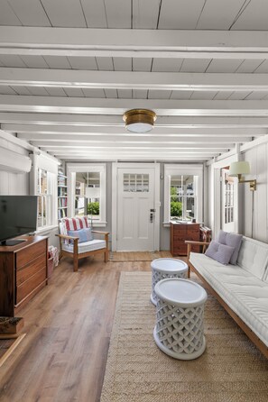 Una habitación con piso de madera, paredes blancas, un sofá blanco, una silla a rayas, un televisor, una lámpara y una puerta con ventanas.