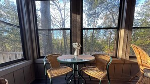 A cozy room with a round table and four wicker chairs, large windows with a view of trees, and a wooden floor.