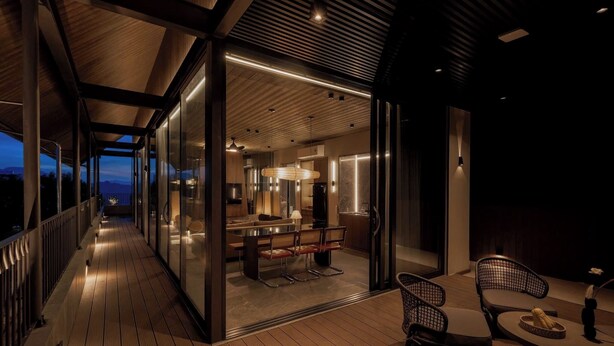 A modern outdoor deck with a wooden floor, glass doors leading to a well-lit interior space, and a seating area with chairs and a table.