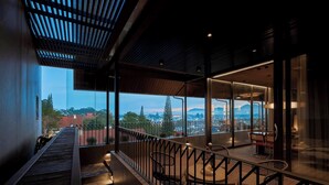 A balcony with a view of the cityscape, featuring a wooden railing, a seating area with chairs, and a staircase leading to the upper level.