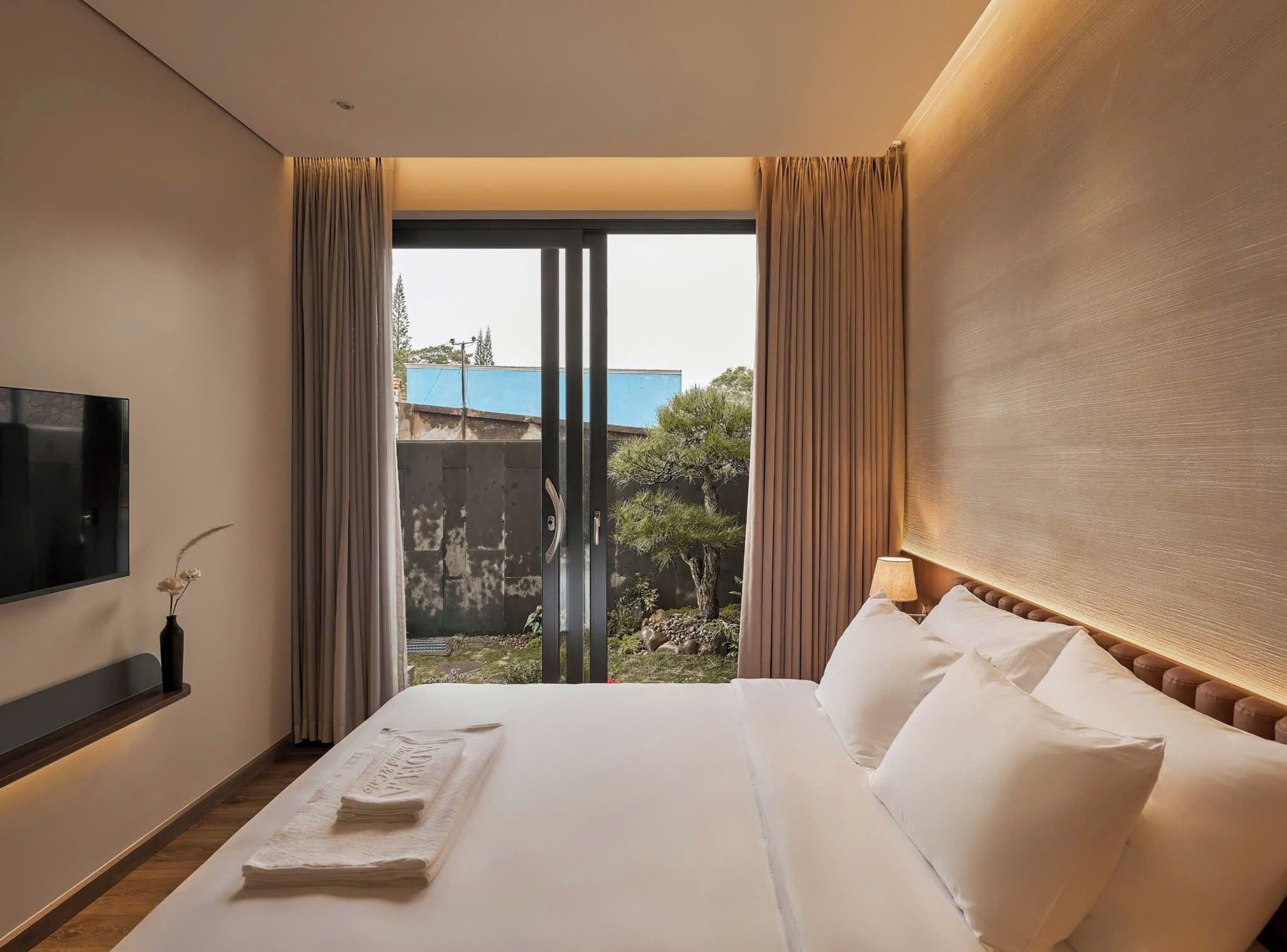A hotel room with a large bed, a flat-screen TV, a bedside table with a lamp, and a view of a building and trees through the window.