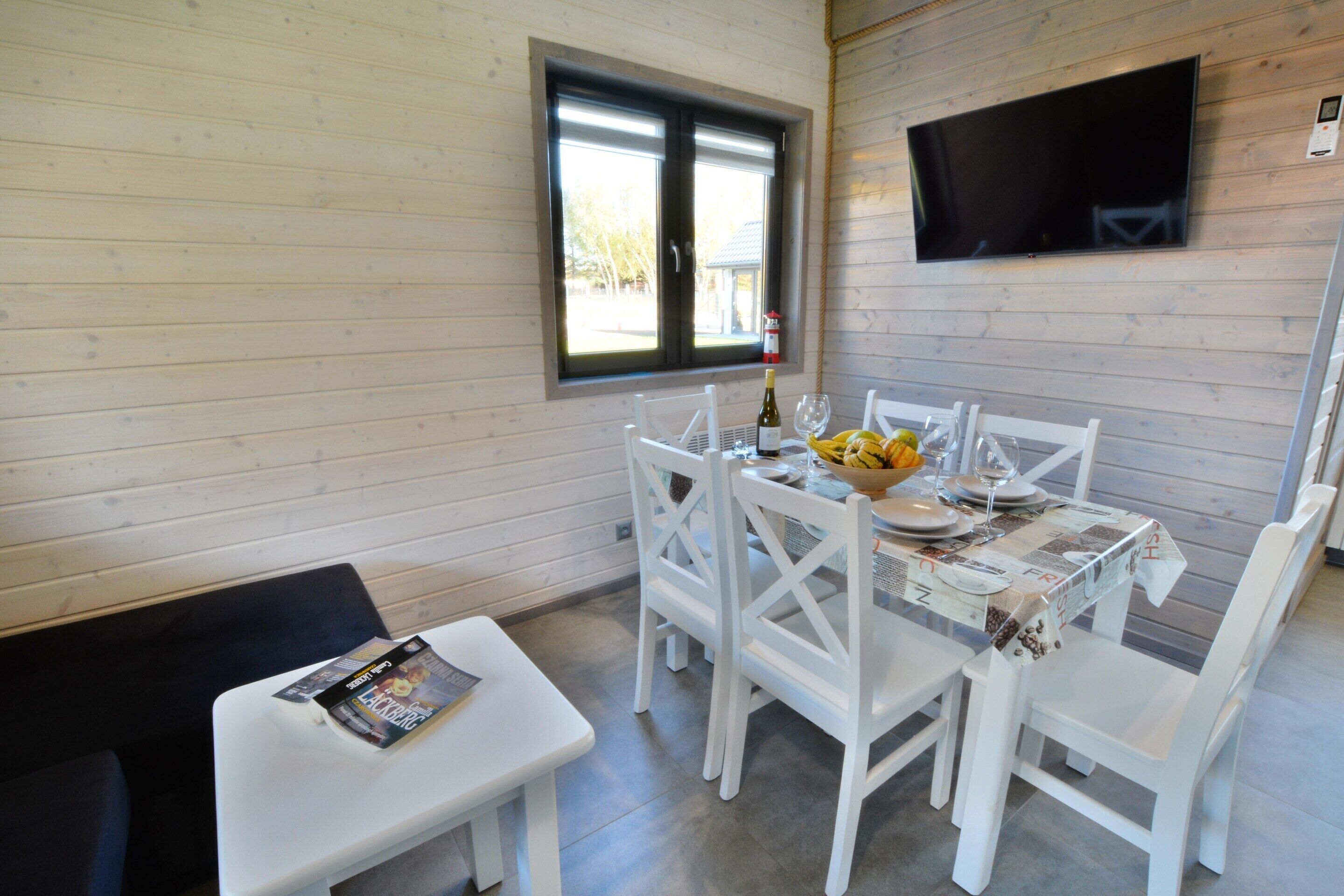 A dining area with a table set for four, white chairs, a small table with a magazine, a window with a view, and a flat-screen TV mounted on the wall.