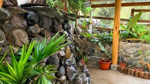 Un mur de pierres avec une petite cascade, entouré de plantes en pot et d’une clôture en bois.