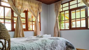 Un lit bien fait, recouvert d’une courtepointe à motifs, une fenêtre donnant sur un paysage verdoyant et des rideaux attachés pour laisser la lumière entrer.