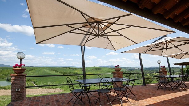 An outdoor patio with brick flooring, metal tables and chairs, beige umbrellas, and flower planters. - Nice Apartment With Swimming Pool (Peccioli)