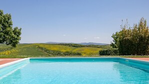 A rectangular swimming pool with clear blue water, surrounded by a red brick deck. The pool is set against a backdrop of green hills and a clear sky. - Nice Apartment With Swimming Pool (Peccioli)