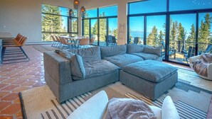 A modern living room with a large sectional sofa, a fireplace, and a view of the forest through large windows.