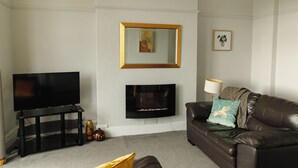 A living room with a flat-screen TV on a black stand, a fireplace, a brown leather sofa with cushions, and a framed picture on the wall. - Seaside Solace: 2BR with Scenic Coastline Views (Llandudno)