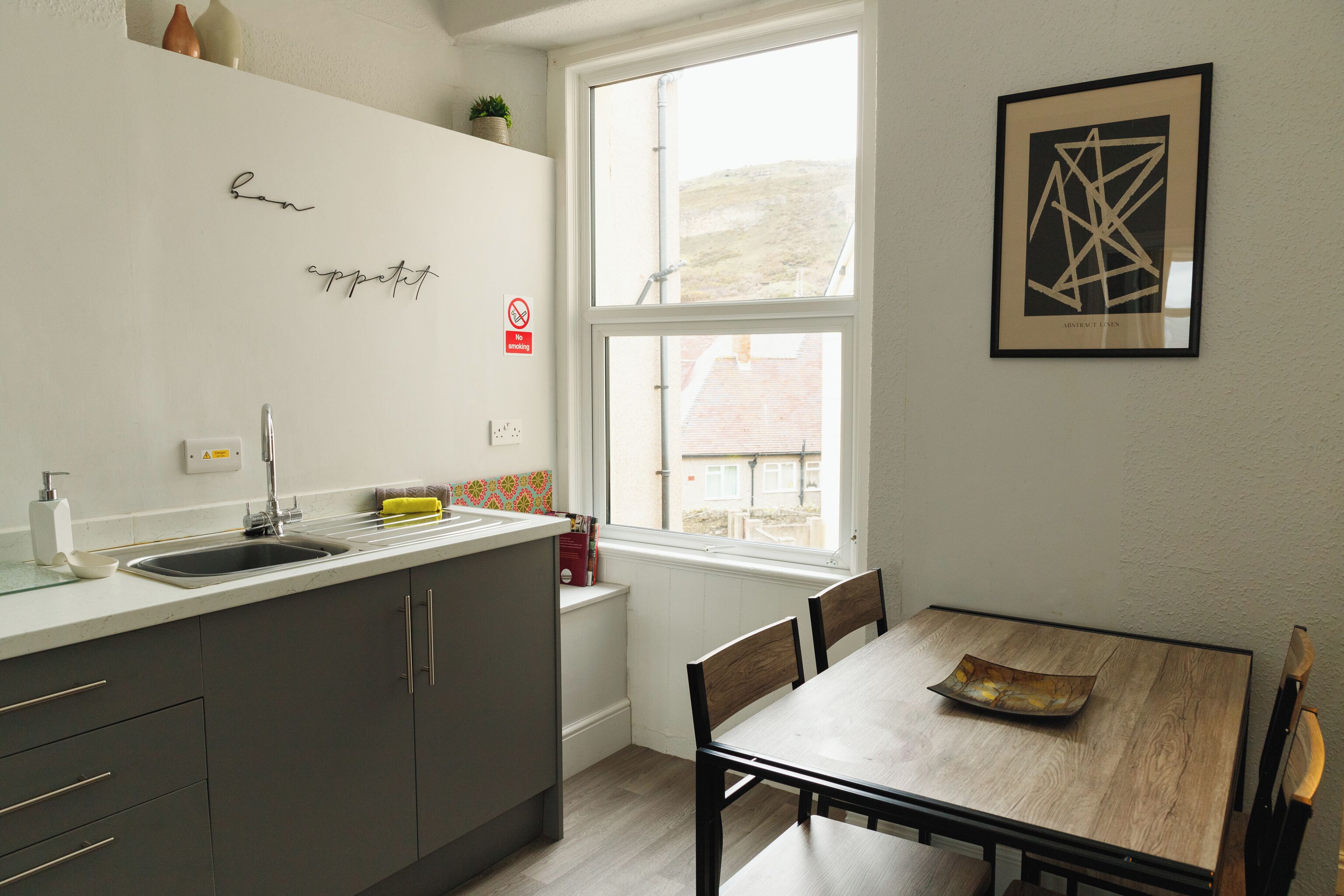 A modern kitchen with a sink, cabinets, and a window. There is a dining table with chairs and a framed picture on the wall.