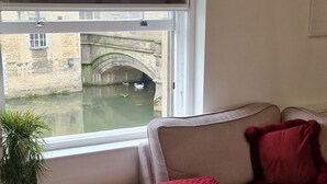 A room with a window overlooking a canal, a sofa with a red blanket, and a wooden floor. - Kingfisher Suite - Two-bed Private Apartment (Stamford)