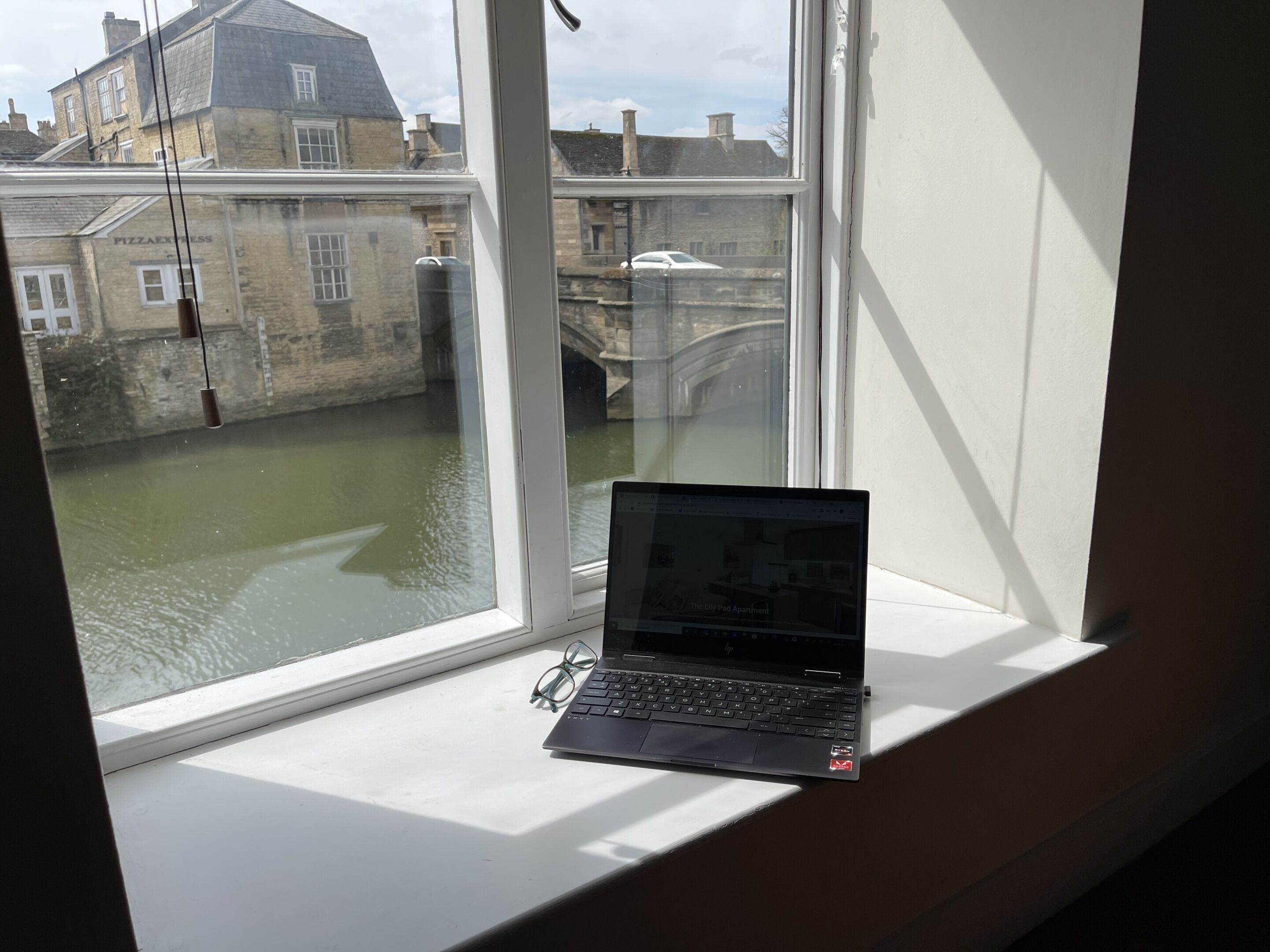 A laptop on a window sill with a view of a river and buildings.