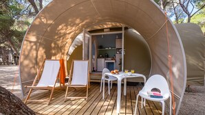 A cozy tent with a wooden deck, two white chairs, a small table, and a kitchenette visible through the entrance. - Premium Tent Lodge for Family (Tarragona)
