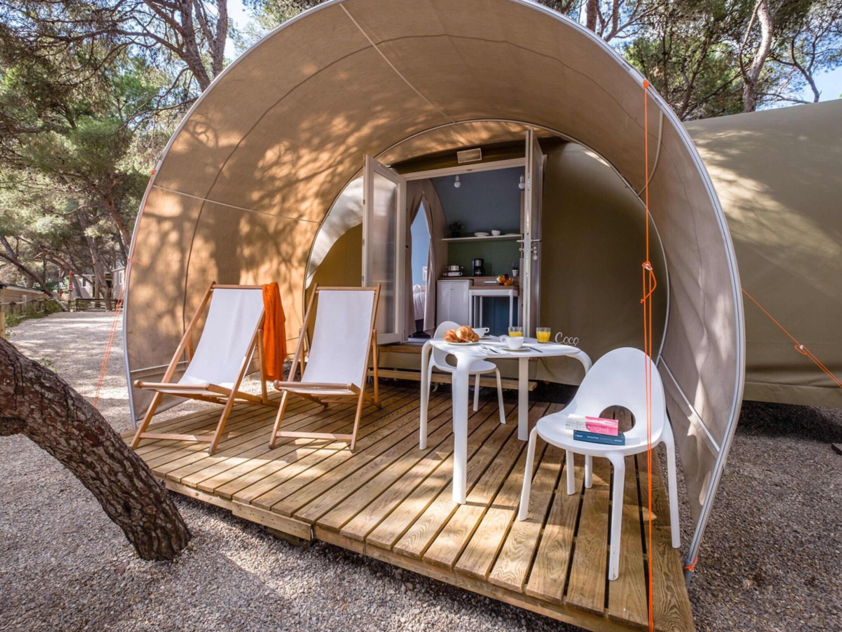 Une tente accueillante, semi-ouverte, dotée d’une terrasse en bois, de deux chaises blanches, d’une petite table et d’une kitchenette visible sur le côté.