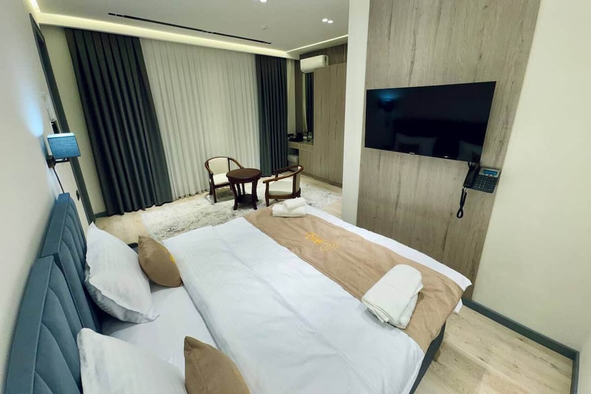 A modern hotel room with a large bed, a flat-screen TV mounted on the wall, a small round table with chairs, and a large window with curtains.