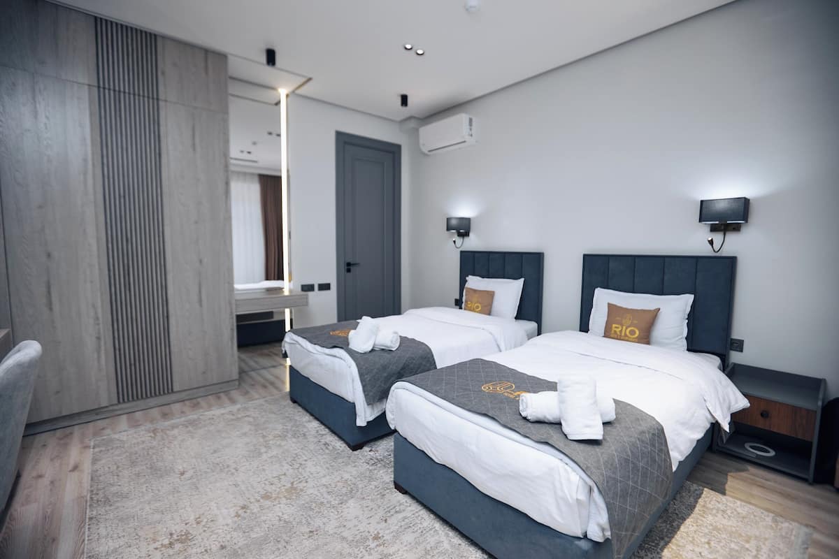 A modern hotel room with two beds, a wooden wardrobe, a mirror, and a bedside table with a lamp.