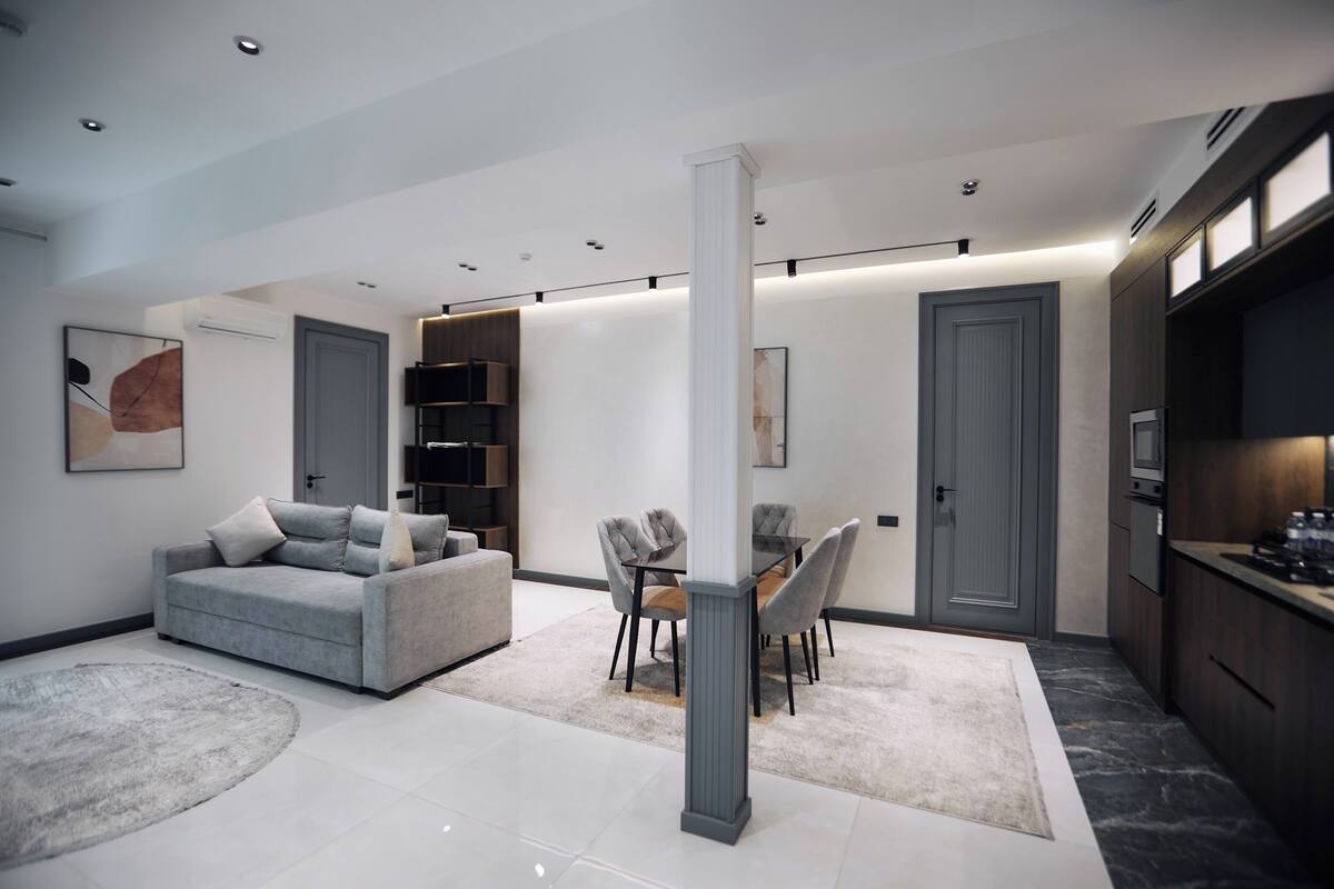 A modern living room with a grey sofa, a dining table with chairs, a large pillar, and a kitchen area with cabinets and a microwave.