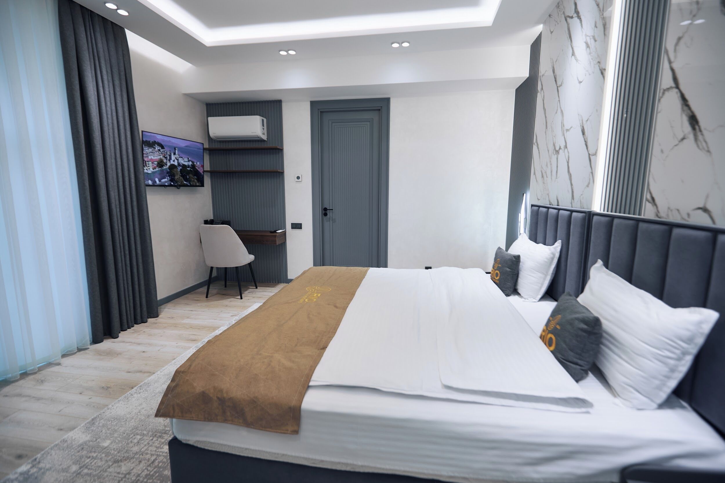 A modern hotel room with a large bed, a desk, a chair, and a television on the wall.
