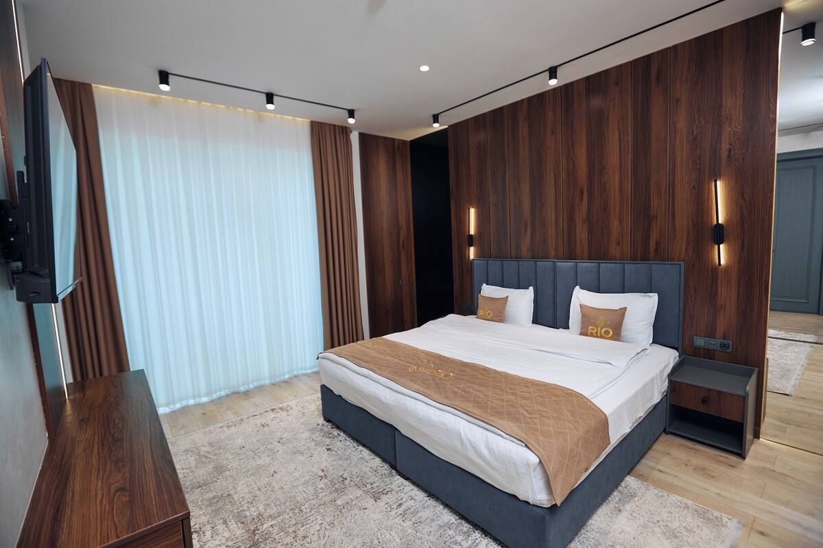 A modern bedroom with a large bed, wooden panel walls, and a flat-screen TV mounted on the wall.