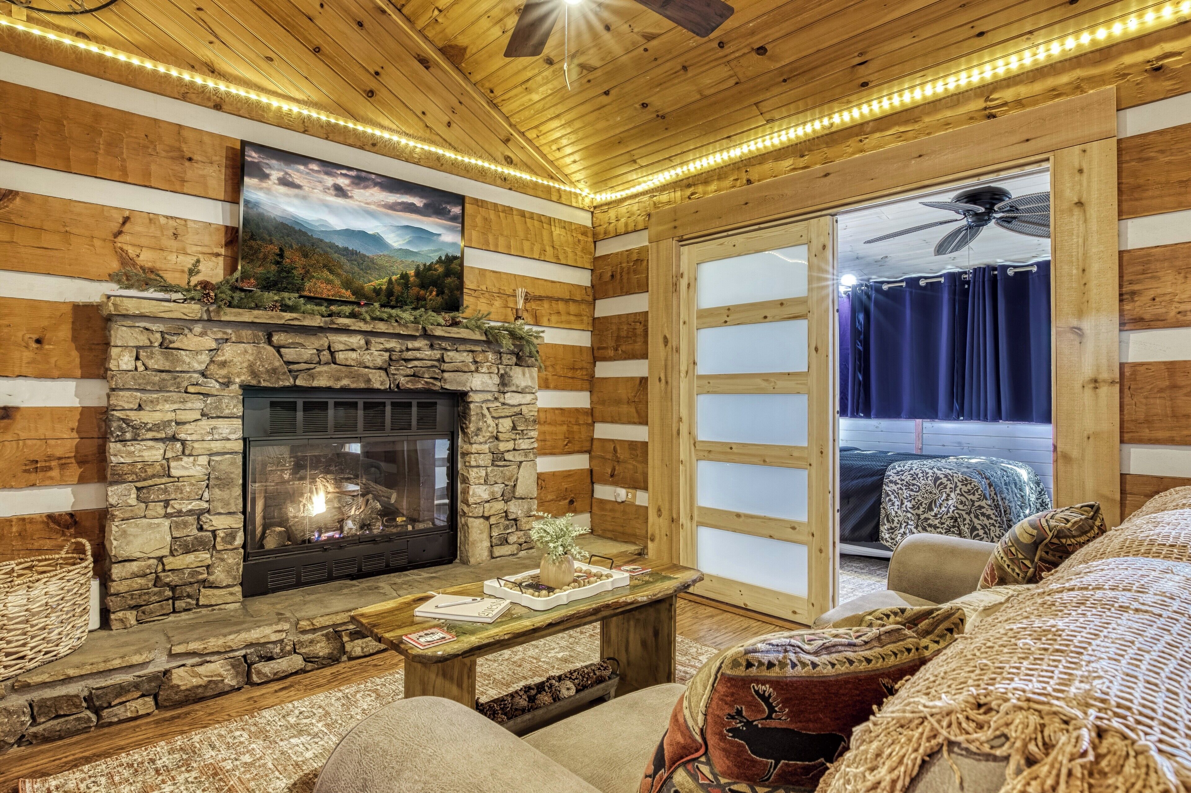 A cozy cabin interior with a stone fireplace, wooden walls, a large window with curtains, and a ceiling fan.