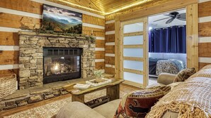 A cozy cabin interior with a stone fireplace, wooden walls, a large window with curtains, and a ceiling fan. - A Timber Hideaway, Cozy Cabin Retreat in the Smokies – Comfort & Fun for All! (Sevierville)