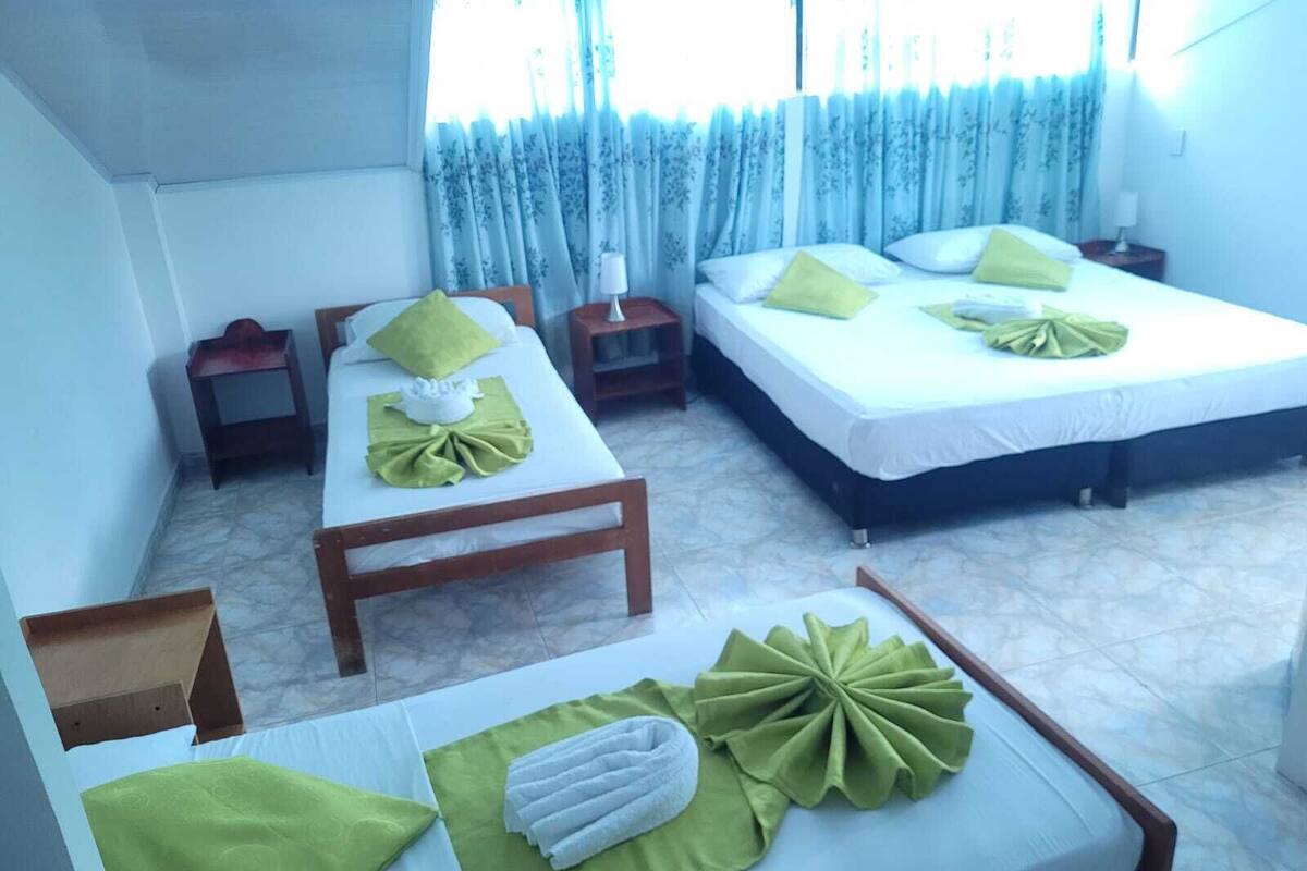 Quarto de hotel com duas camas, cada uma com toalhas e almofadas verdes, uma pequena mesa de madeira e uma mesa de cabeceira com luminária.