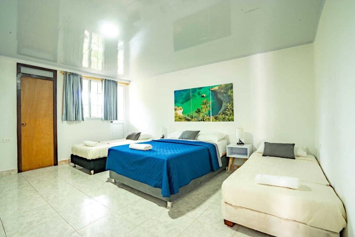 Quarto de hotel com duas camas, uma janela grande com cortinas, um quadro na parede e chão revestido de azulejos.