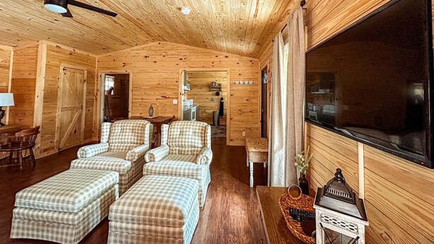 Un accogliente interno di una baita in legno, con un grande televisore, poltrone a quadri, una ventola a soffitto in legno e un'area pranzo con tavolo e sedie.