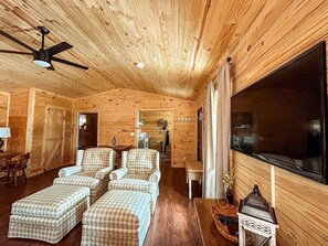 A cozy log cabin interior with a large TV, checkered armchairs, a wooden ceiling fan, and a dining area with a table and chairs.