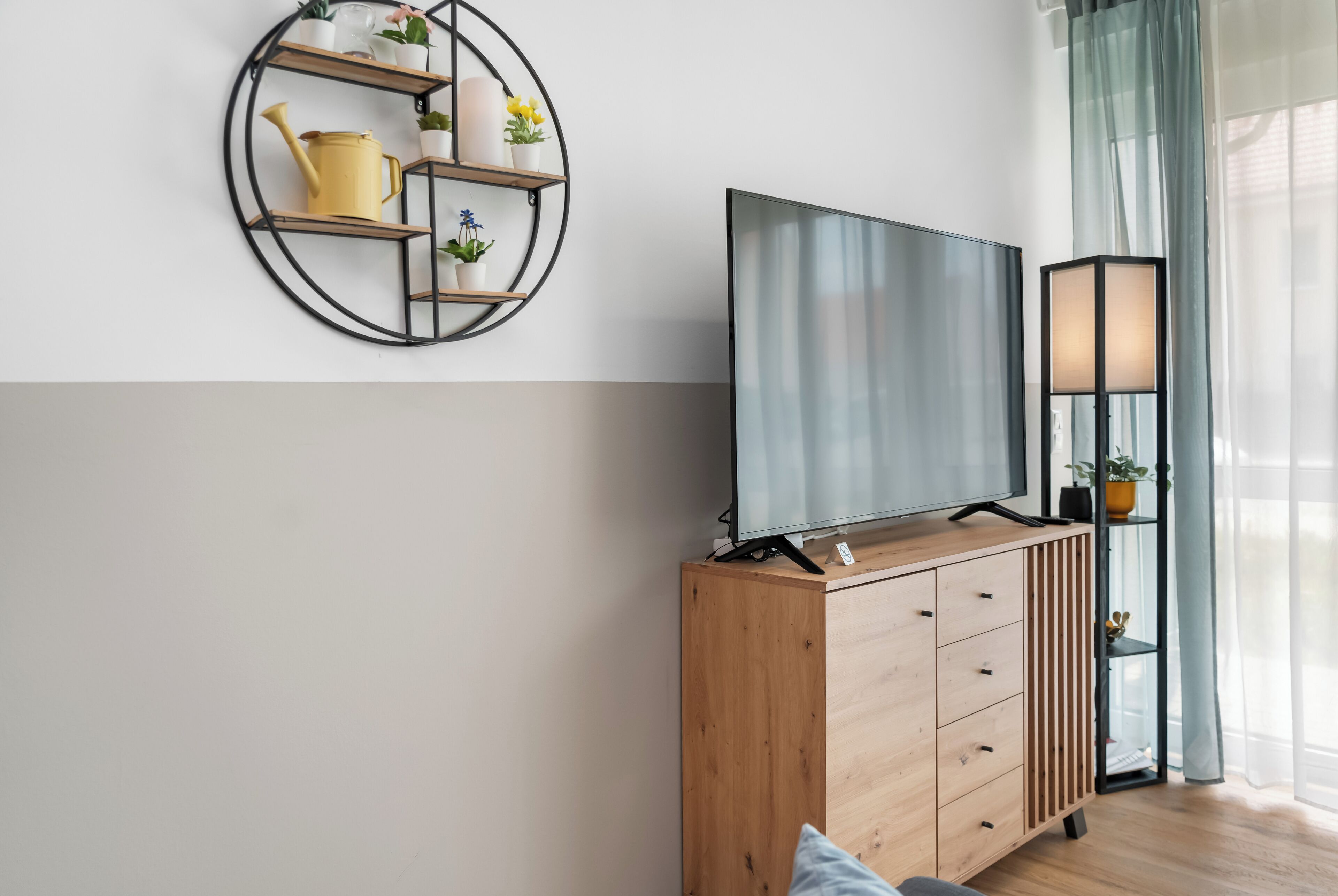 A modern living room with a wooden cabinet, a flat-screen TV, a round wall shelf with plants, and a floor lamp.