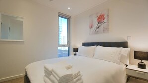 A hotel room with a large bed, a nightstand with a lamp, a window with a view of buildings, and a painting on the wall. - Amyra Residences on Rowlands Place - Unit 1505 (Adelaide)