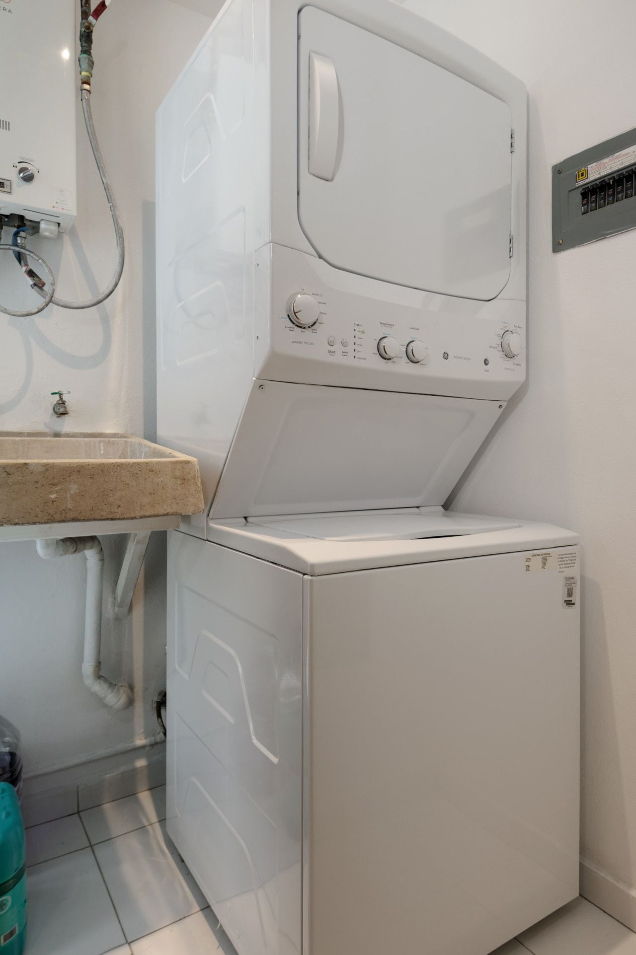 Un appareil compact combinant lave-linge et sèche-linge, doté d’un panneau de commande, d’une arriv ée d’eau et d’un panneau électrique fixé au mur.