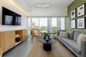 A modern living room with a flat-screen TV, a sofa, a coffee table, and a large window offering a city view.