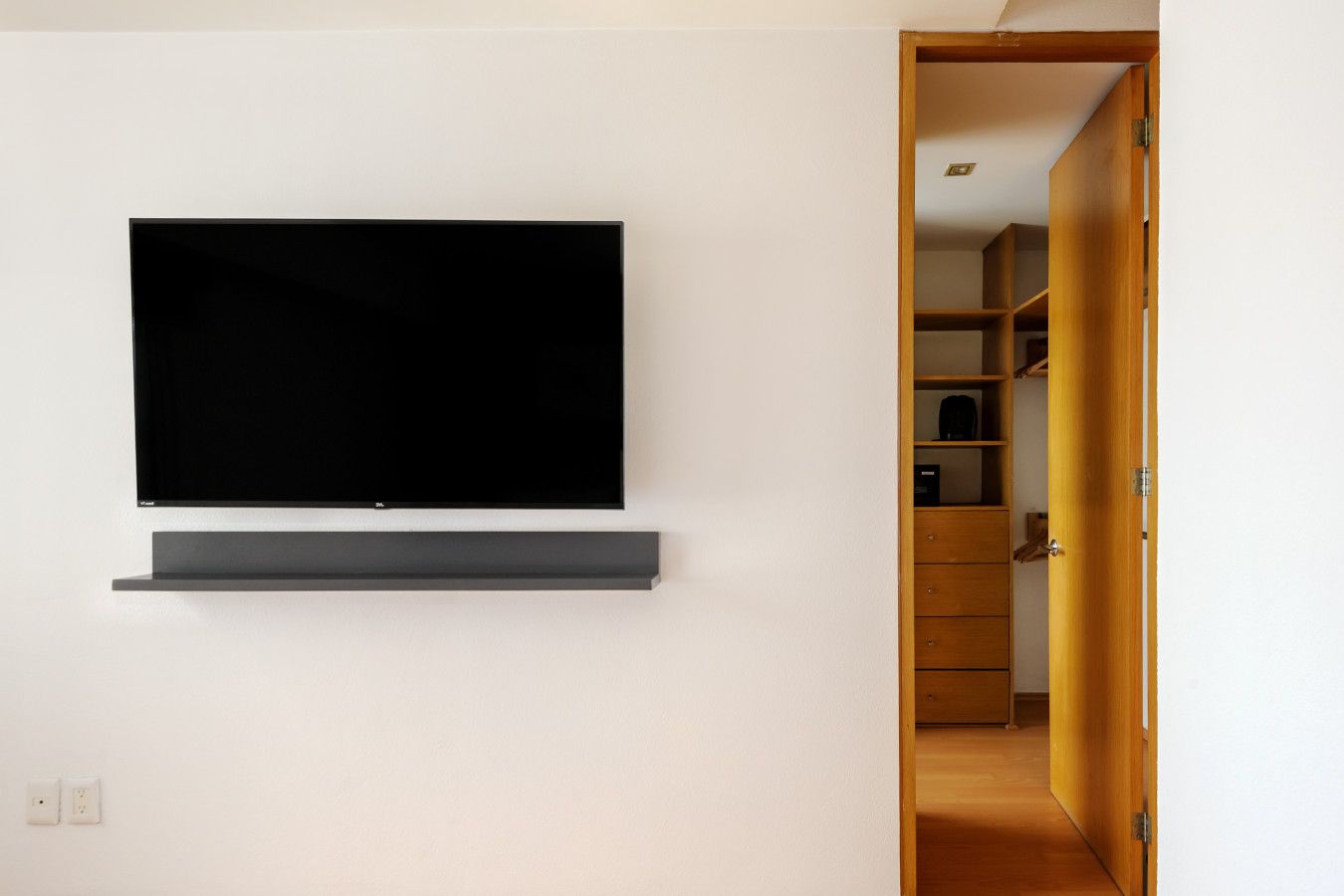 A modern room with a flat-screen TV mounted on the wall, a wooden door leading to a closet, and a white wall.