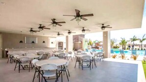 An outdoor dining area with multiple tables and chairs, ceiling fans, and a view of a swimming pool. - Casa de las Olas (La Paz)